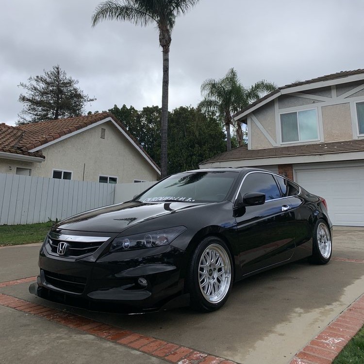 Black coupe with silver wheels parked in a driveway under a cloudy sky